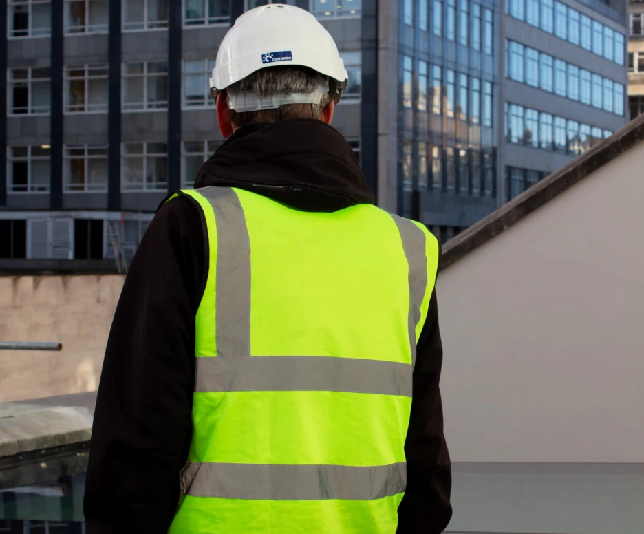A person wearing a high-visibility vest and a white hard hat stands with their back to the camera, looking at a modern cityscape featuring glass buildings.