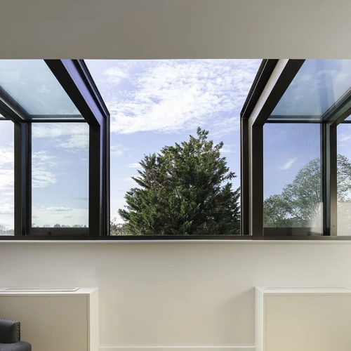 Large, modern windows open outward, revealing a clear blue sky with scattered clouds and a green tree outside. The interior features a minimalist design with light wooden flooring and white wall-mounted furniture.