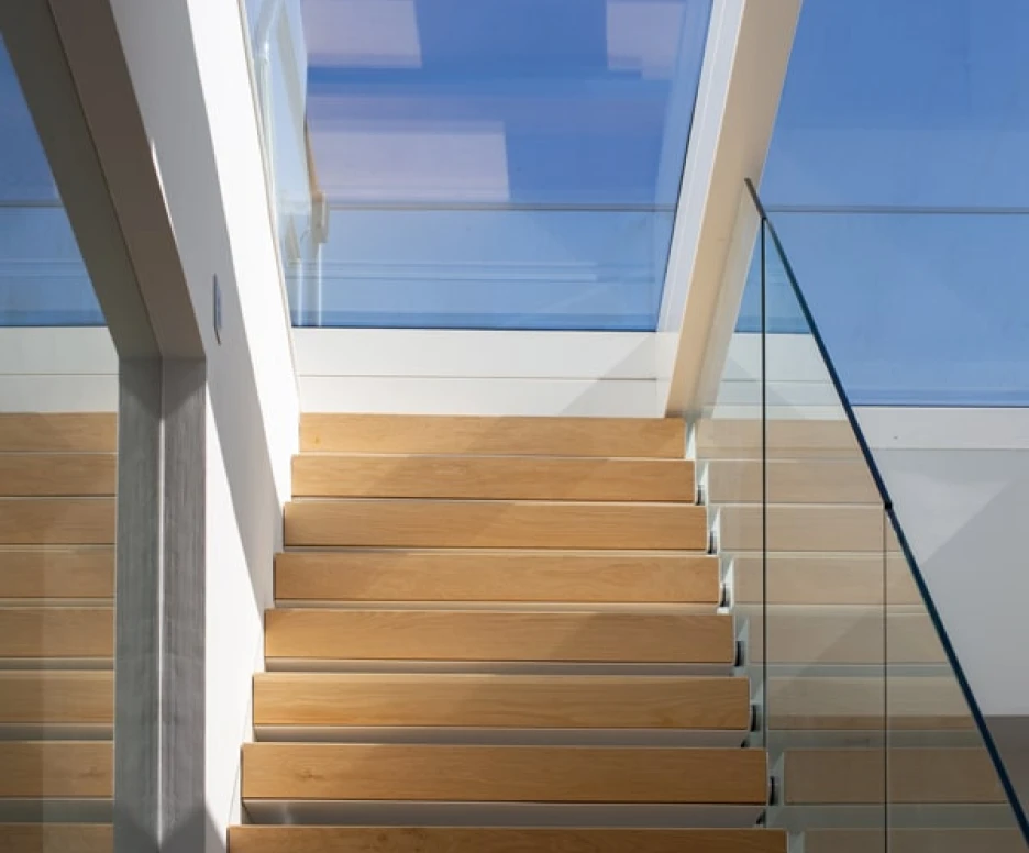 Wooden staircase leading upwards, illuminated by natural light filtering through a glass roof. The stairwell features sleek glass railings and minimalist architecture.