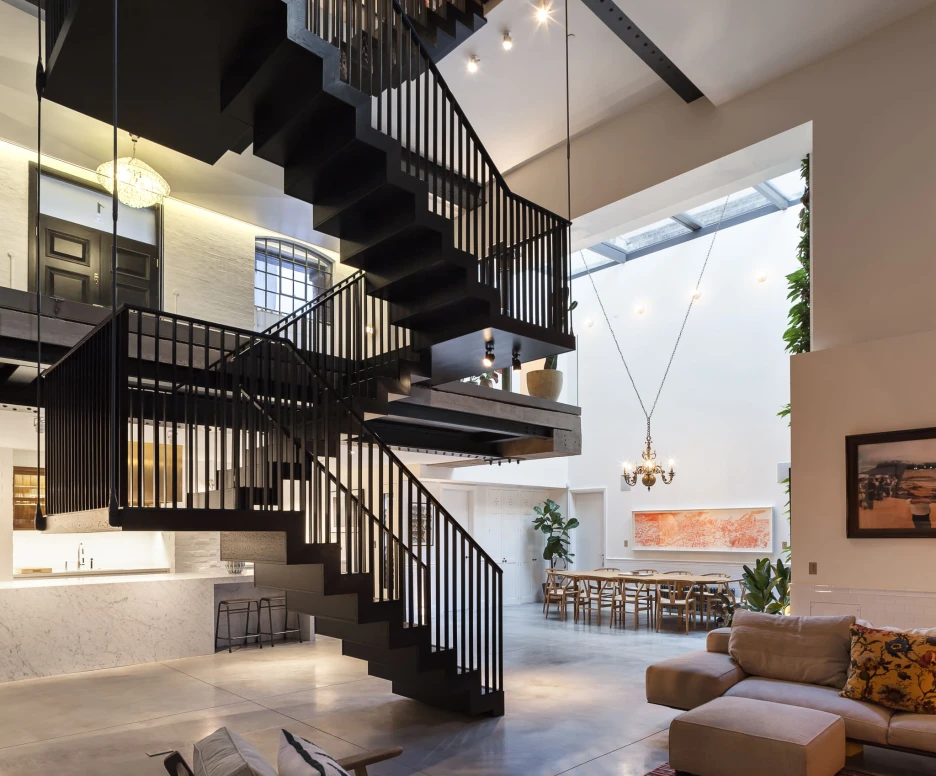 A modern interior featuring a sleek black spiral staircase with railing. The space has high ceilings, large windows, and an open layout. A cozy living area with a sectional sofa and colorful rug is visible, along with dining space and greenery in the background. Stylish lighting fixtures hang from the ceiling.