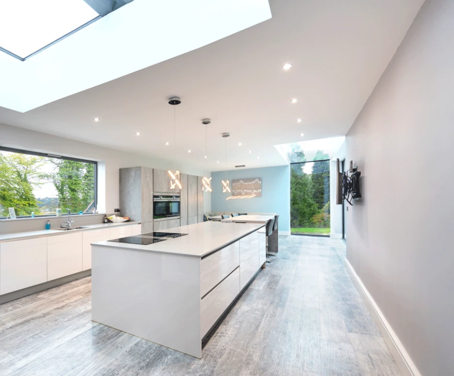 A modern kitchen featuring a large island with a cooktop, sleek white cabinetry, and large windows that bring in natural light. The space has a minimalist design with a light wood floor and a view of greenery outside.
