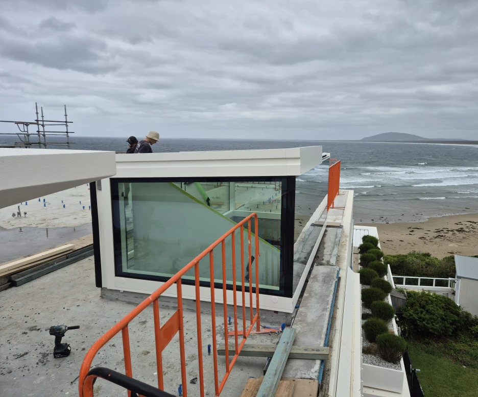 A modern glass-enclosed structure sits on the edge of a building overlooking a beach and ocean. Two people stand on the rooftop, observing the view. The sky is overcast and the waves are visible in the distance.