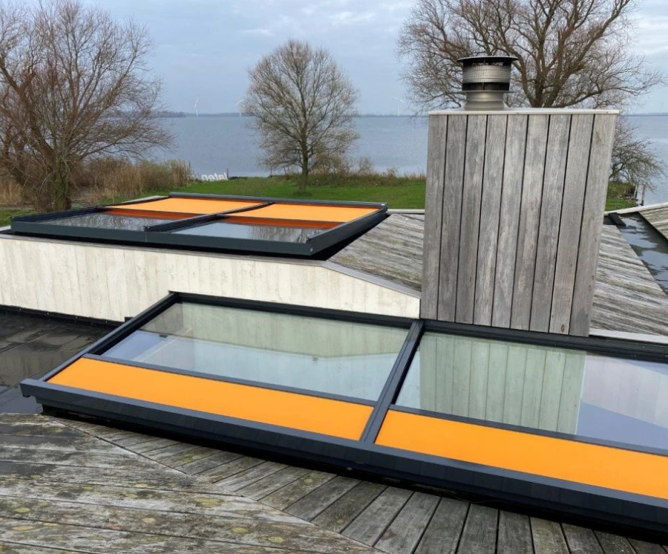 A rooftop view featuring two large skylights with orange frames. In the background, a body of water and trees can be seen, with a chimney on the side.