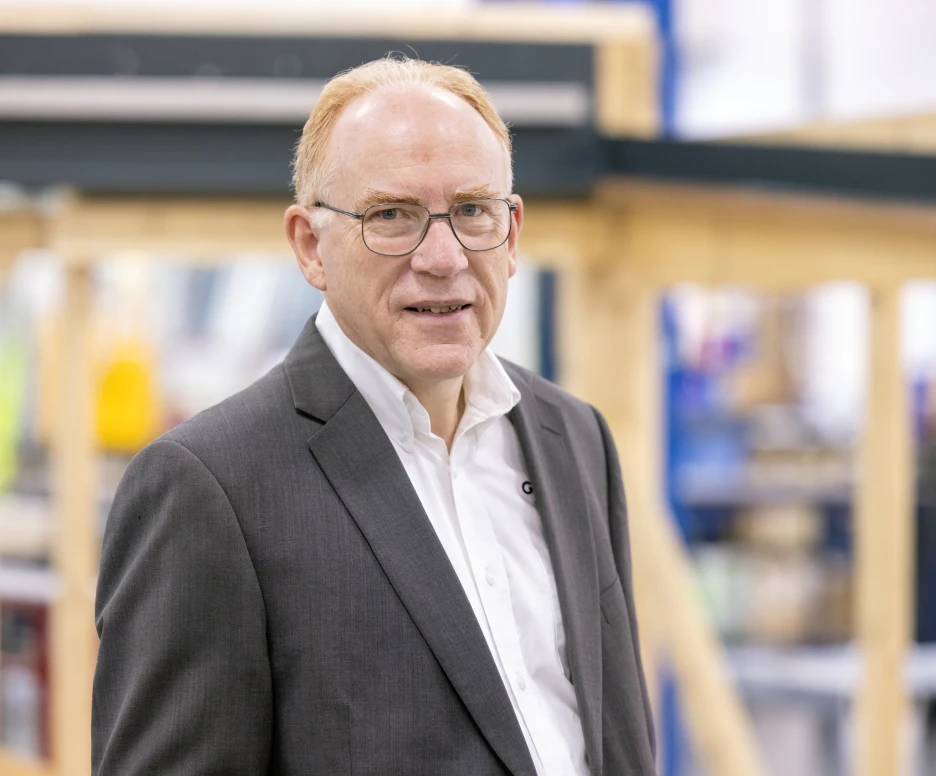 A middle-aged man with glasses wearing a dark suit and white shirt stands confidently in a workshop setting filled with wooden structures.