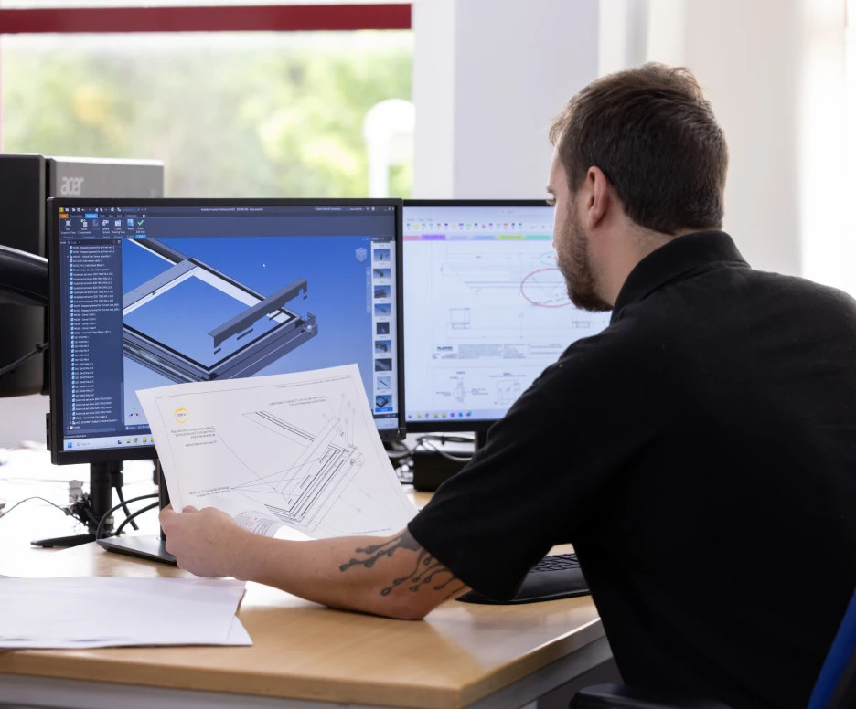 A person sitting at a desk, reviewing engineering plans while looking at a computer screen displaying a 3D model. The workspace includes multiple monitors, paperwork, and a bright background with windows.