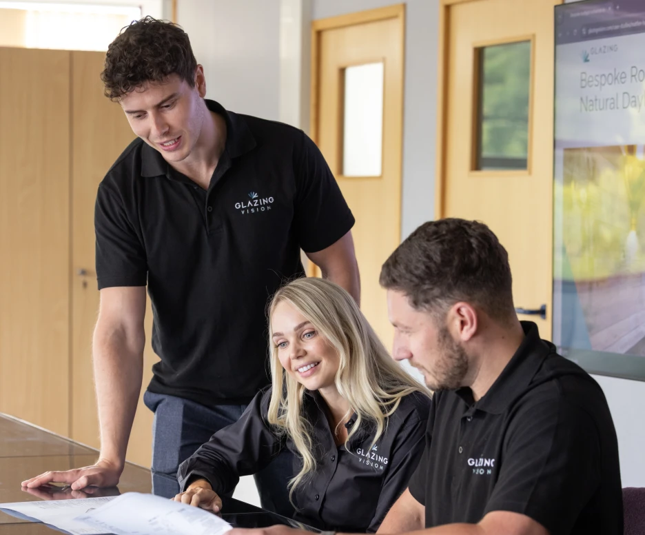 Three people collaborating on a project at a table. A man stands to the left, while a woman and another man sit at the table, reviewing documents. All three are wearing black polo shirts with a logo. A screen in the background displays information.