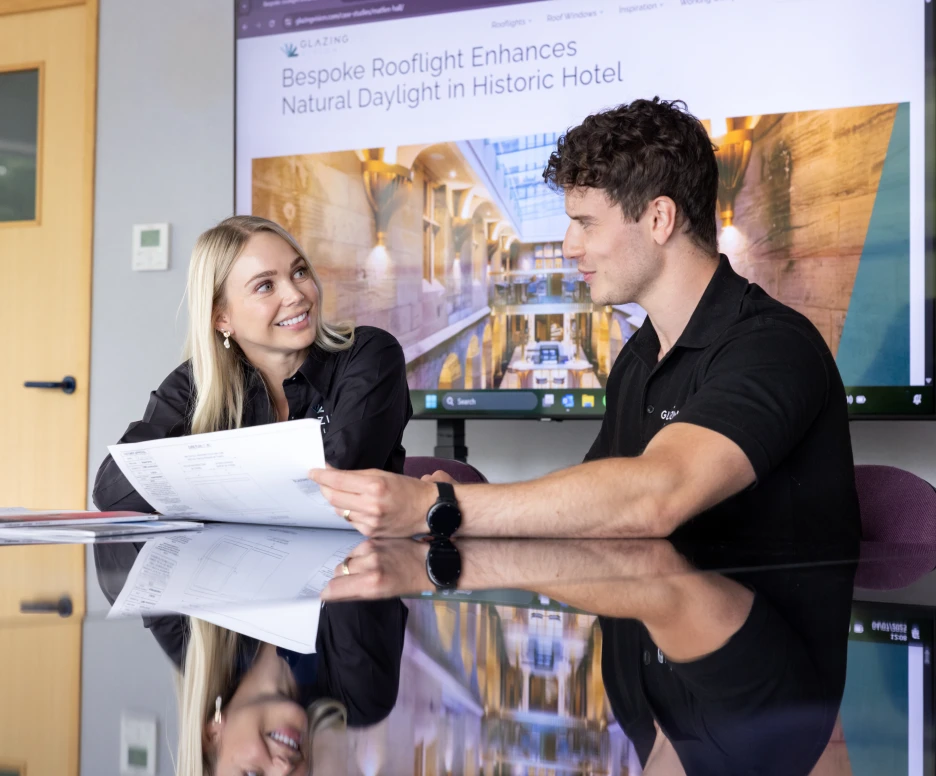 Two people are engaged in a discussion at a conference table, reviewing plans and documents. A large display in the background features an article about enhancing natural daylight in a historic hotel. Both individuals are wearing black shirts with the company's logo.