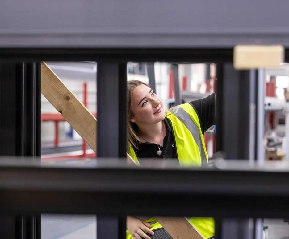 A woman in a bright yellow safety vest is working in a manufacturing setting, smiling as she adjusts a piece of wood in a framed structure. The background shows a workshop environment with tools and equipment.