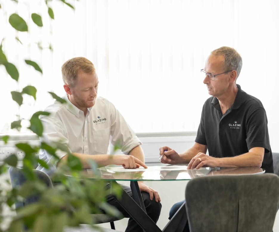 Two men engaged in a discussion at a glass table in a modern office setting. One man points at a document while the other takes notes. Green foliage is visible in the foreground.