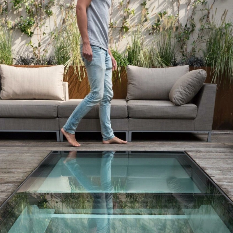 A person walks barefoot over a glass floor section in a modern outdoor space, surrounded by stylish seating and greenery.