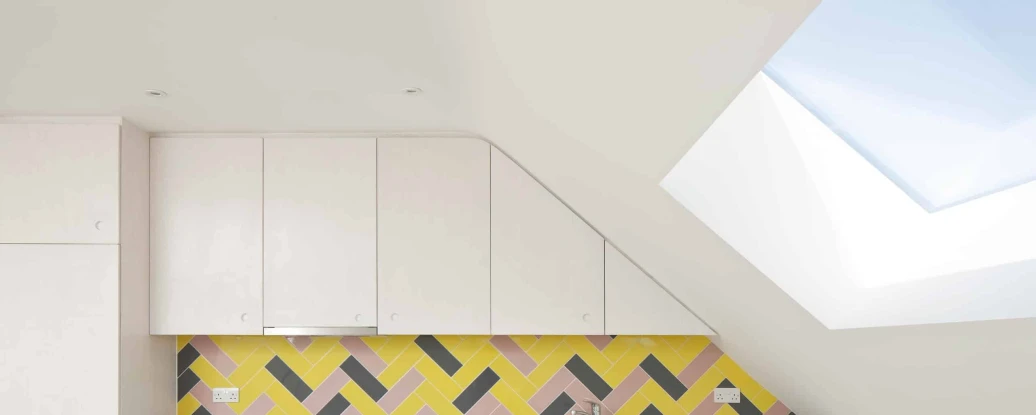 Modern kitchen featuring white cabinets and a colorful herringbone-patterned backsplash in yellow, pink, and gray. A stainless steel stove sits against the wall beneath a skylight that adds natural light to the space.