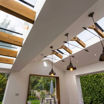 A modern room with a white ceiling featuring wooden beams and skylights. Decorative pendant lights hang from the ceiling, illuminating the space. In the background, a glass door opens to a garden, surrounded by green foliage.