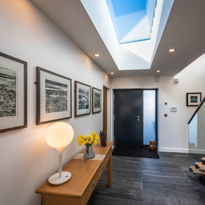 A modern hallway featuring a sleek wooden console table with a lamp and a vase of yellow flowers. Framed black-and-white artwork hangs on the walls, and a large skylight allows natural light to illuminate the space. A dark door leads outside, and a small rug is placed at the entrance.