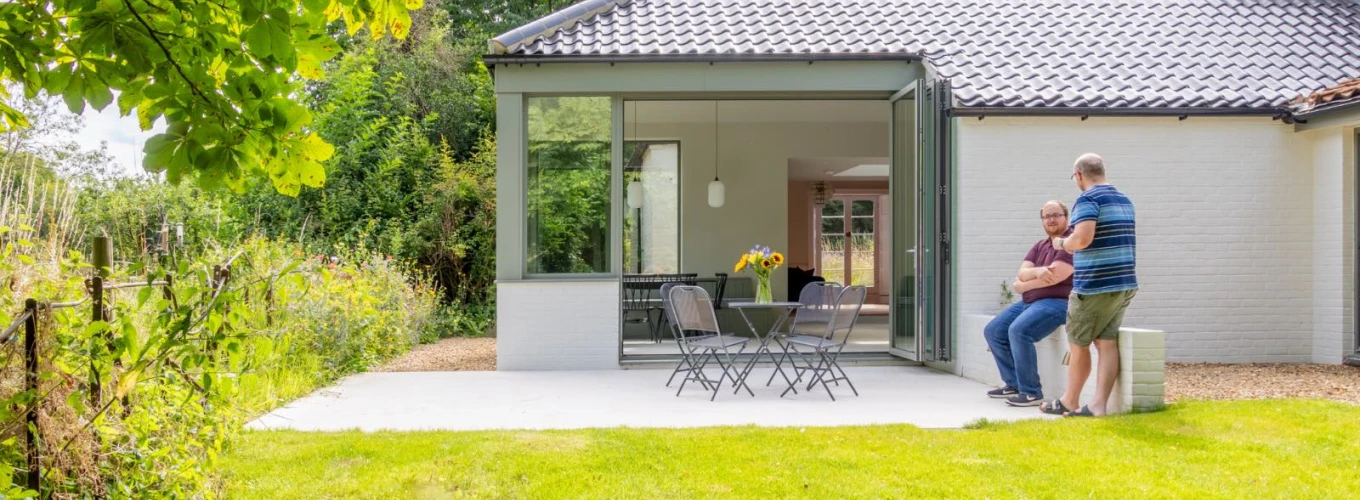 A modern home with a glass-walled extension opens to a lawn area, where two men are seated on a patio. Sunflowers are visible on the table, and lush greenery surrounds the property.