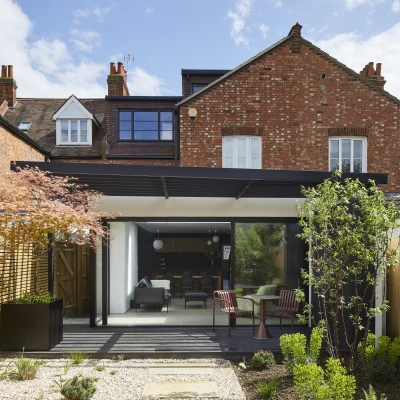 A modern backyard view of a home featuring a sleek glass extension with a wooden deck. Surrounded by greenery and a mix of landscaping, the house has red brick walls and a contemporary black roof. Blue skies with a few clouds enhance the atmosphere.