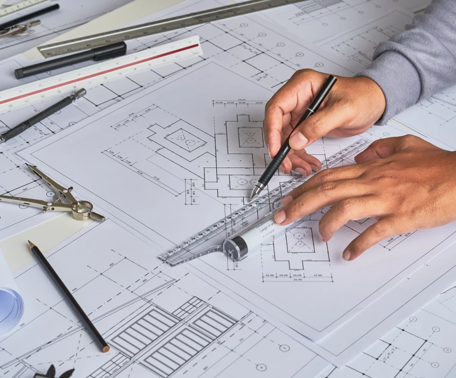 Hands using a pencil and triangular ruler to measure and draw on architectural blueprints spread across a table. Various drafting tools, including a compass and pens, are scattered alongside the plans.