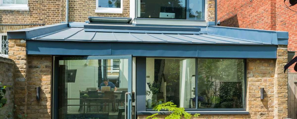A modern single-story extension of a brick house features a glass wall and a blue metal roof. An outdoor table with chairs sits in front of the extension, surrounded by greenery.