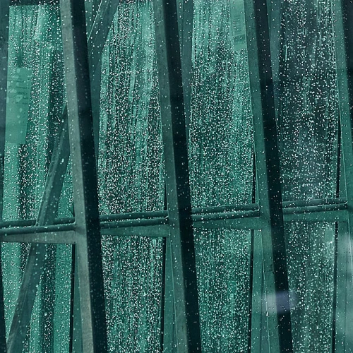 Raindrops on a large glass window, creating a blurred, textured pattern with greenish tones and geometric lines from the window frame.