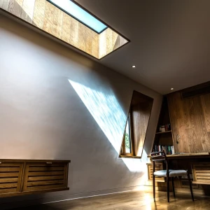 A modern study with wooden furniture, featuring a large skylight that casts a dramatic shadow on the wall. A wooden desk and chair are positioned near a window, with bookshelves visible in the background.