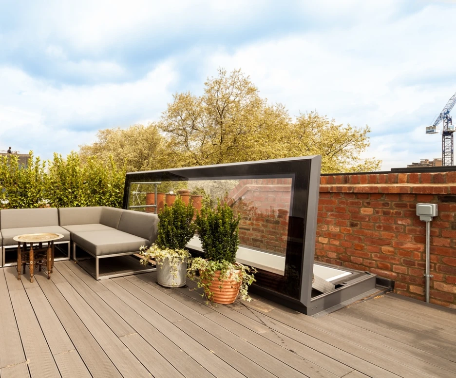 A stylish rooftop patio with gray seating, a round table, and potted plants. The space is surrounded by greenery and features a modern glass partition. Brick walls and a blue sky create a serene atmosphere.