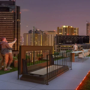 A rooftop gathering at dusk features people enjoying the scenic city skyline. Some are taking selfies, while others are seated at tables with lights strung above. A fire pit is visible, surrounded by greenery, adding to the cozy atmosphere.