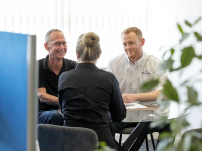 Two men are sitting at a glass table, engaging in a discussion with a woman who has her back to the camera. The setting features bright indoor lighting and plants in the background. The men appear to be smiling and interacting positively.