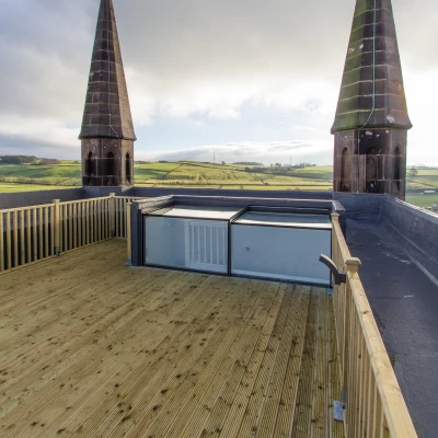 A rooftop view featuring two tall, pointed spires against a cloudy sky. Below, there's a wooden deck and a glass structure, with rolling green hills in the distance.