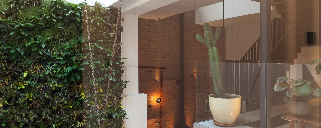 Interior view of a modern space featuring a vertical garden wall, large skylight, and minimalist decor. A staircase is visible in the background alongside stylish furnishings, while a chandelier hangs above a dining table.