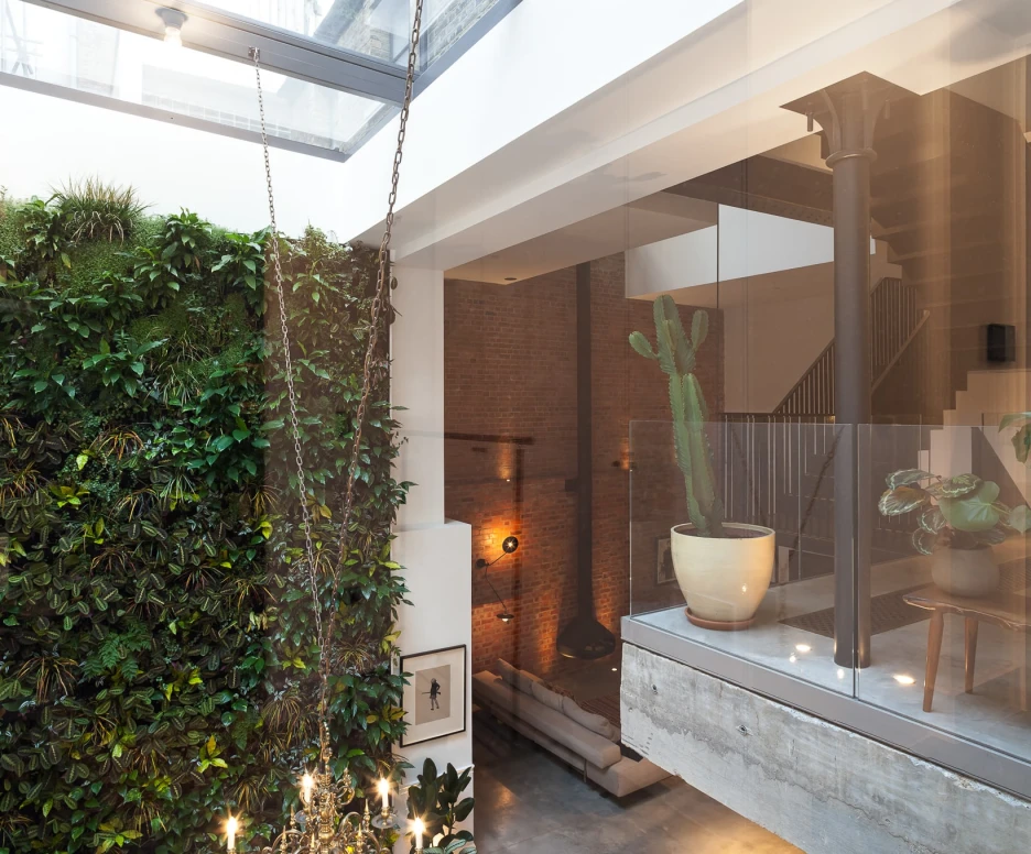 Interior view of a modern space featuring a vertical garden wall, large skylight, and minimalist decor. A staircase is visible in the background alongside stylish furnishings, while a chandelier hangs above a dining table.