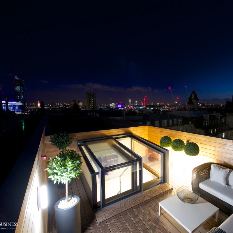 A terrace which overlooks London. On the terrace you can see a white curved sofa which is looking at the lit up box skylight which leads to terrace.