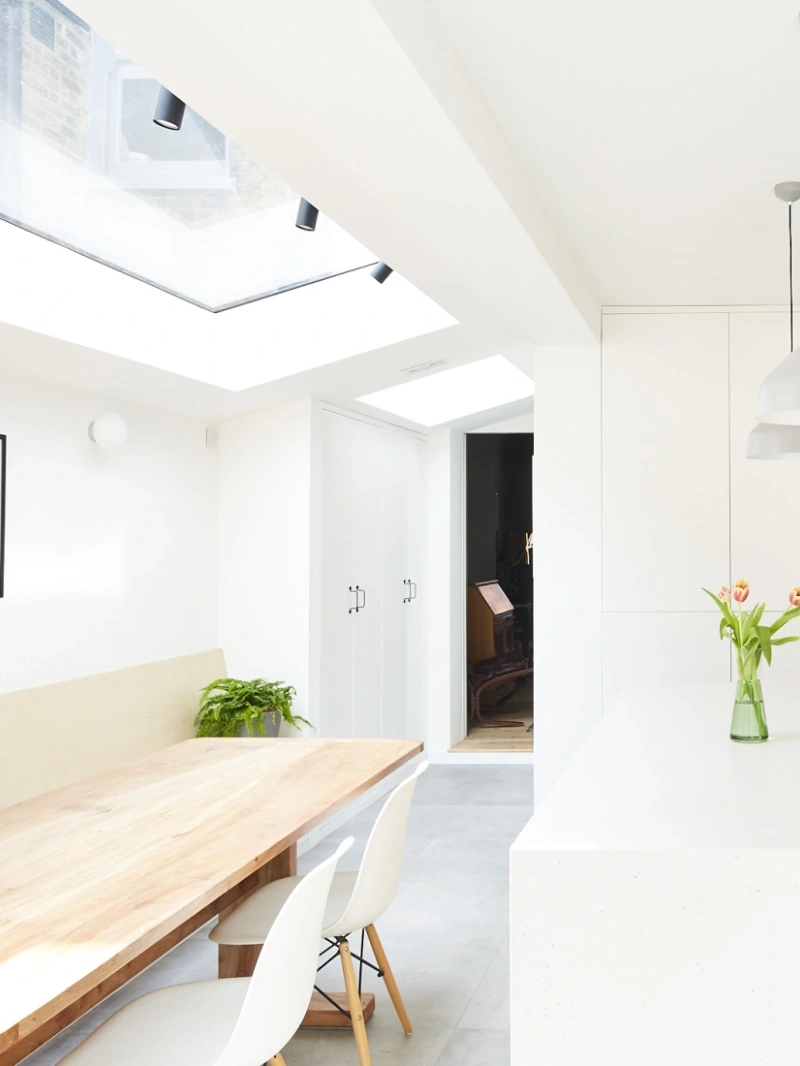 A bright, modern kitchen and dining area featuring a natural wood dining table, white chairs, and a vase of tulips. Large skylights provide ample natural light, highlighting the minimalist decor with white walls and cabinetry.