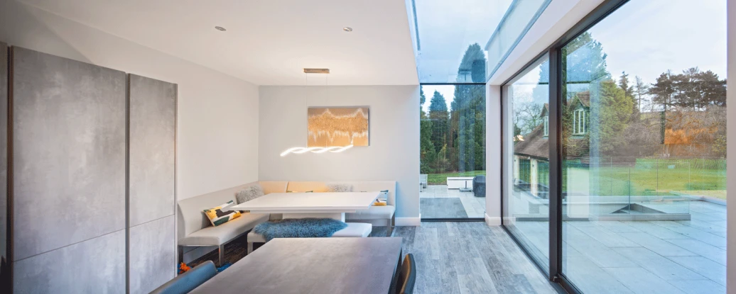 A modern dining area featuring a sleek table and a built-in bench with cushions. Large glass walls provide a view of the outdoor landscape, while a stylish light fixture hangs above the table. Natural light floods the space, enhancing the contemporary design.