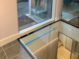 Large modern window with a view of a patio and garden. A reflection of a person is visible on the glass. The interior features a sleek design with a glass floor and grey tiles.