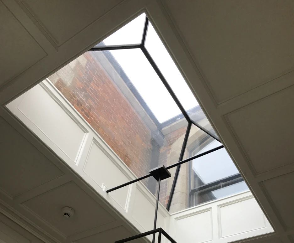 A modern interior featuring a skylight with a geometric black frame. A stylish black and glass chandelier hangs below the skylight, illuminating the space. Elegant white paneling surrounds the area.