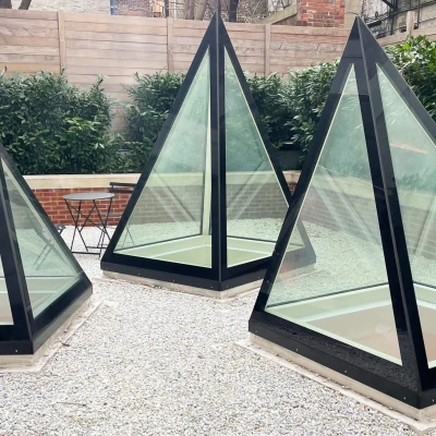 Three glass pyramids with black frames are situated in a gravel area surrounded by greenery and a low brick wall. A small table and chair are visible in the background.