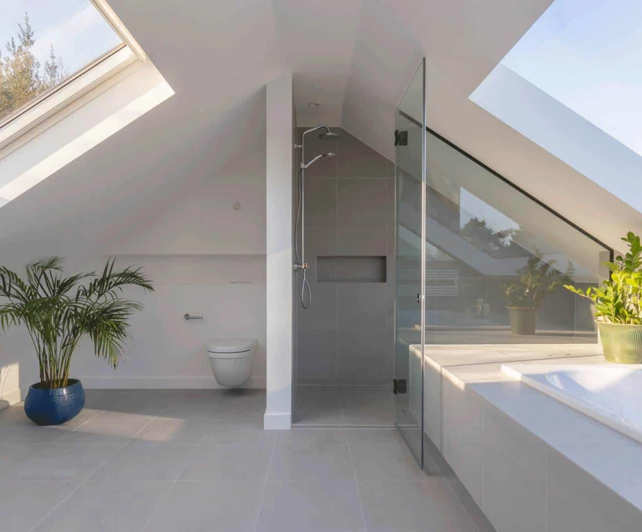 A modern bathroom with sloped ceilings featuring a glass shower, a white toilet, and a bathtub. Natural light streams in through a skylight, and a potted plant adds a touch of greenery. The floor is tiled in light gray.