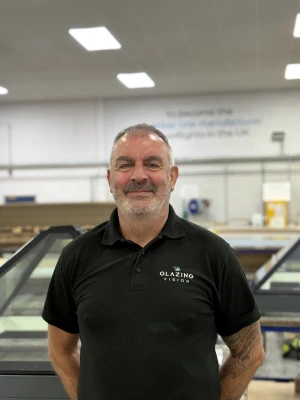 A smiling man with short gray hair stands in a well-lit workshop. He is wearing a black polo shirt with a logo that says "Glazing Vision." In the background, there are glass structures and equipment typical of a manufacturing environment.