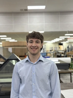 A young man with curly hair smiles while wearing a light blue, polka-dotted shirt. He stands in a bright workshop filled with various tools and equipment in the background.