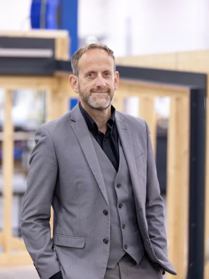 A well-dressed man in a gray suit stands confidently with his hands in his pockets against a backdrop of a manufacturing or workshop environment. He has short hair and a beard, and looks directly at the camera.