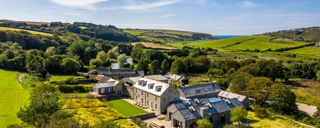 A picturesque countryside scene featuring a large stone house with multiple wings, surrounded by lush green fields and trees. Rolling hills extend into the distance, and the coastline is visible under a bright blue sky with wispy clouds.