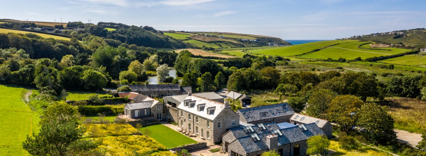 A picturesque countryside scene featuring a large stone house with multiple wings, surrounded by lush green fields and trees. Rolling hills extend into the distance, and the coastline is visible under a bright blue sky with wispy clouds.