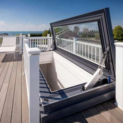 A rooftop deck featuring a large, open hatch that leads below. The surrounding area is adorned with white railings and two lounge chairs, with a scenic view of the sea and blue sky in the background.