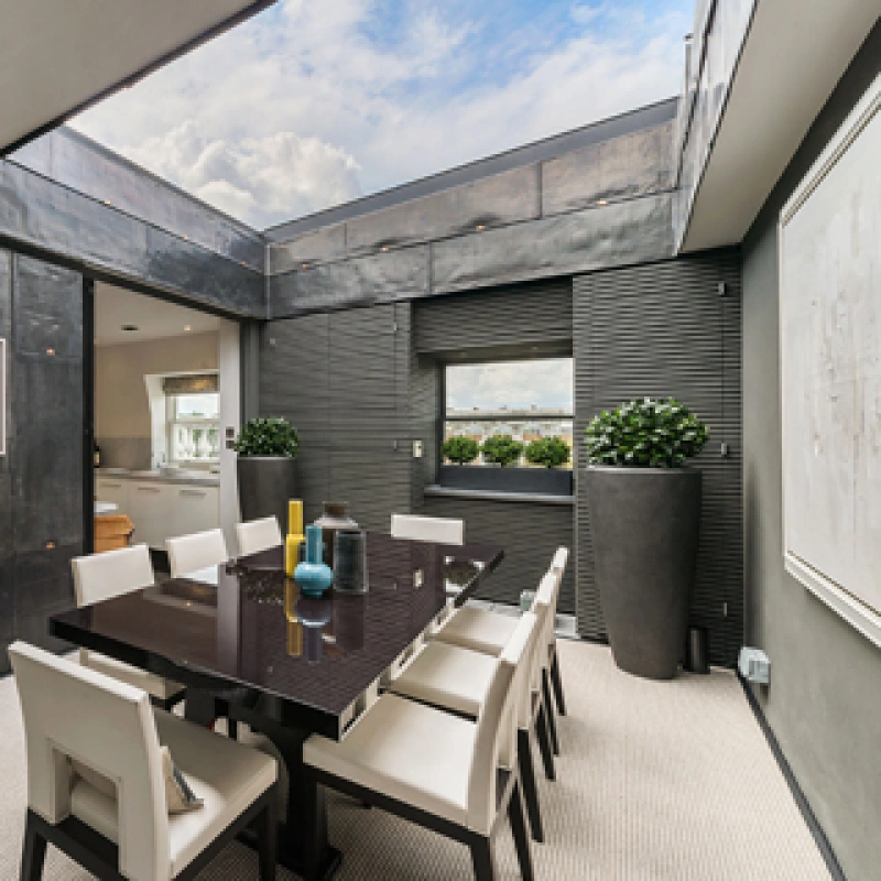 A modern dining room featuring a large dark wooden table surrounded by white chairs. The room has sleek dark walls with a skylight, allowing natural light to enter. Stylish decor includes potted plants and contemporary artwork on the walls.