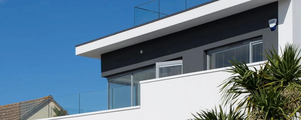 A modern two-story house with a flat roof and a glass railing on the upper balcony. The exterior features a combination of white and gray walls, surrounded by palm trees against a clear blue sky.