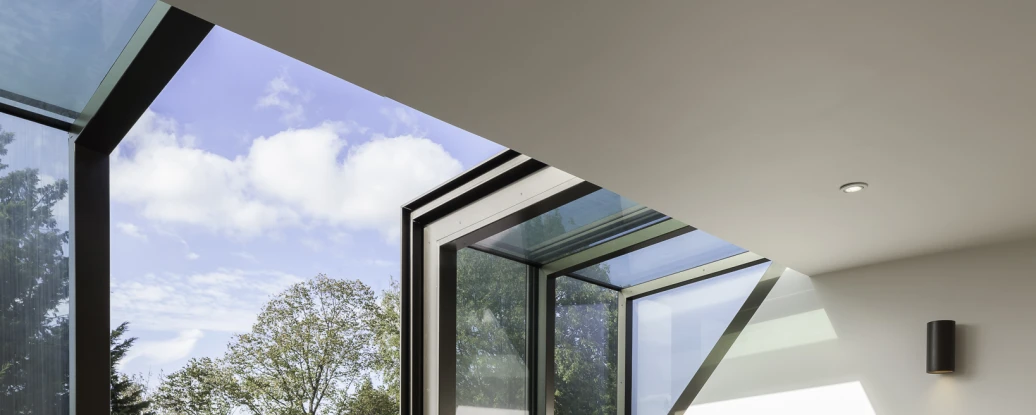Two gray upholstered chairs are positioned in a well-lit room with large modern windows showcasing a blue sky and trees outside. Light wooden flooring complements the space, which has minimalist decor and built-in shelving.