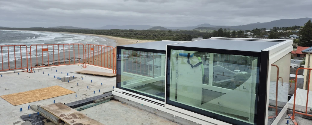 A modern glass structure under construction on a rooftop, overlooking a beach with turbulent gray skies and distant mountains. Safety barriers surround the area, and wooden planks are placed nearby.