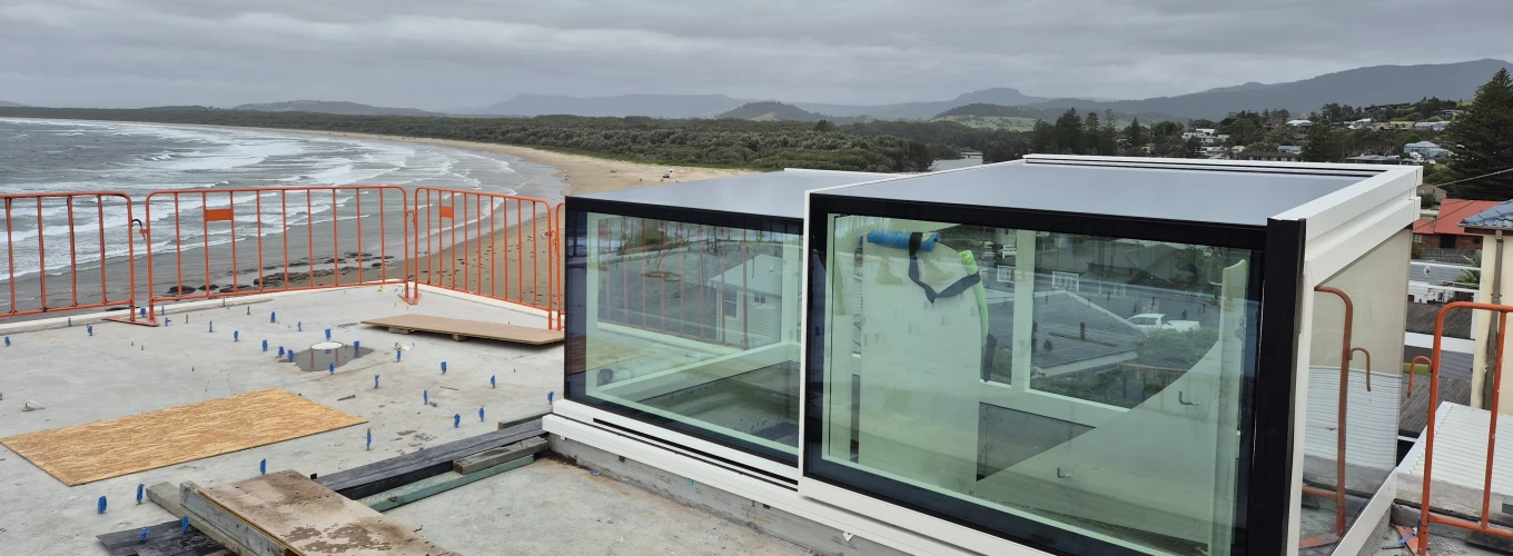 A modern glass structure under construction on a rooftop, overlooking a beach with turbulent gray skies and distant mountains. Safety barriers surround the area, and wooden planks are placed nearby.