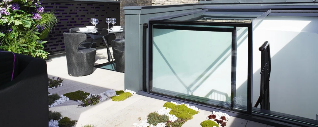 A modern rooftop terrace features a glass canopy, a table set for two with black chairs, and greenery among stone and wooden elements. In the background, a multi-story brick building is visible against a clear blue sky.