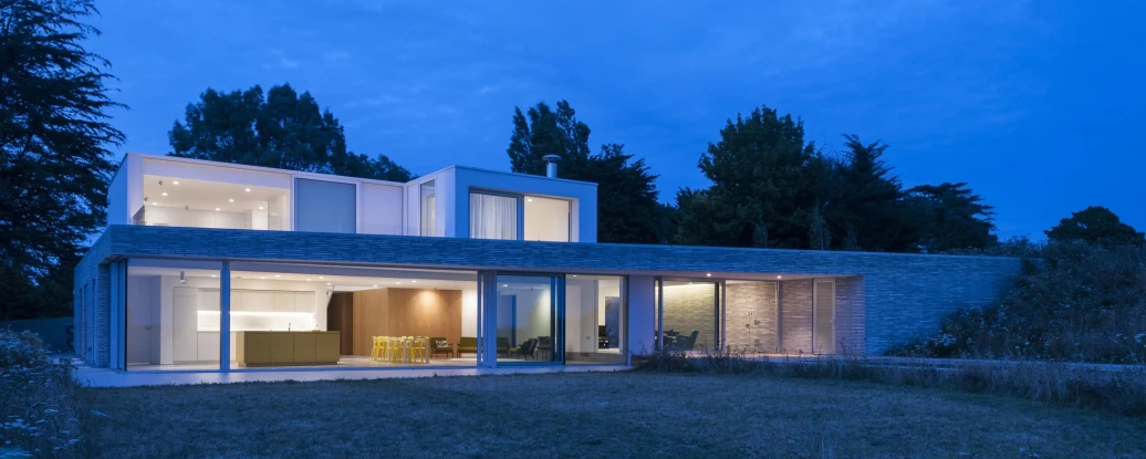 A modern two-story house with large glass windows and a flat roof, set in a landscaped yard under a twilight sky. The light from the interior creates a warm ambiance, and trees frame the background.