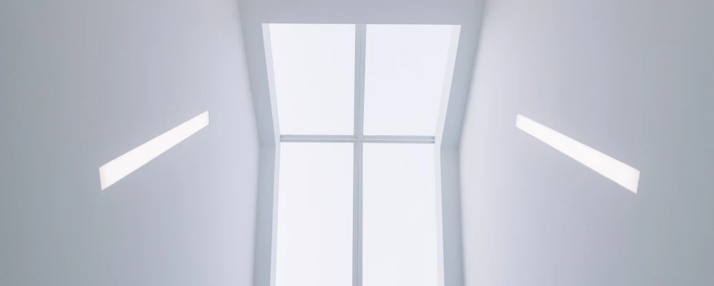 A view looking upward at a bright, minimalist stairwell. The ceiling is mostly white, featuring recessed lighting and a large rectangular window that lets in natural light.